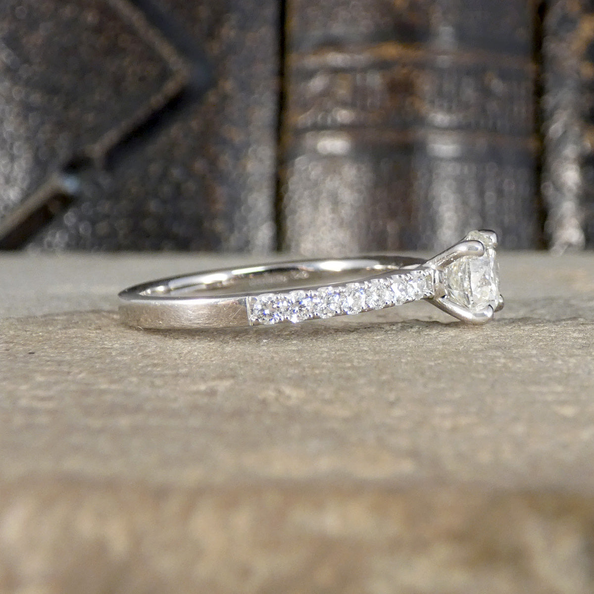 A silver-toned diamond ring with a diamond set band and a round-cut center stone, resting on a textured stone surface with a blurred dark background.