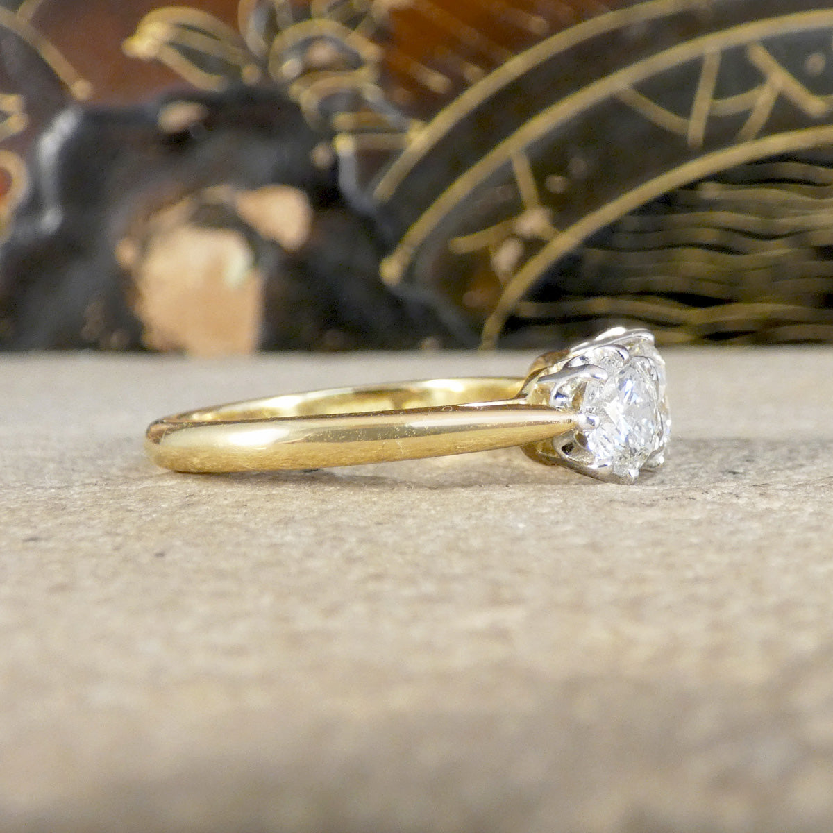 A yellow gold engagement ring with a round brilliant-cut diamond, resting on a textured beige stone surface with a blurred decorative background.