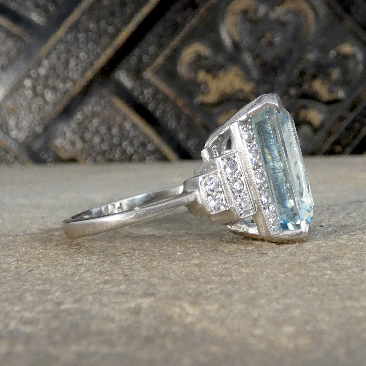 A platium ring featuring a large rectangular light blue gemstone with smaller clear stones on the shoulders, resting on a textured stone surface with a dark decorative background.