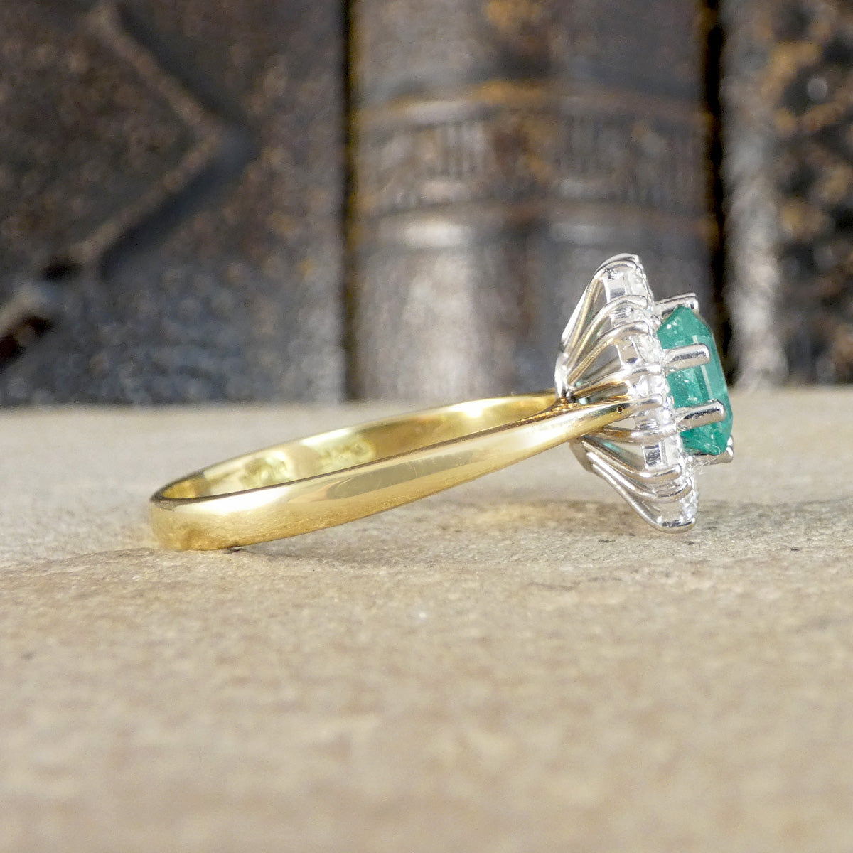 A yellow gold ring with a silver-toned halo setting and a green emerald-cut center stone, resting on a textured beige surface with a blurred dark background.