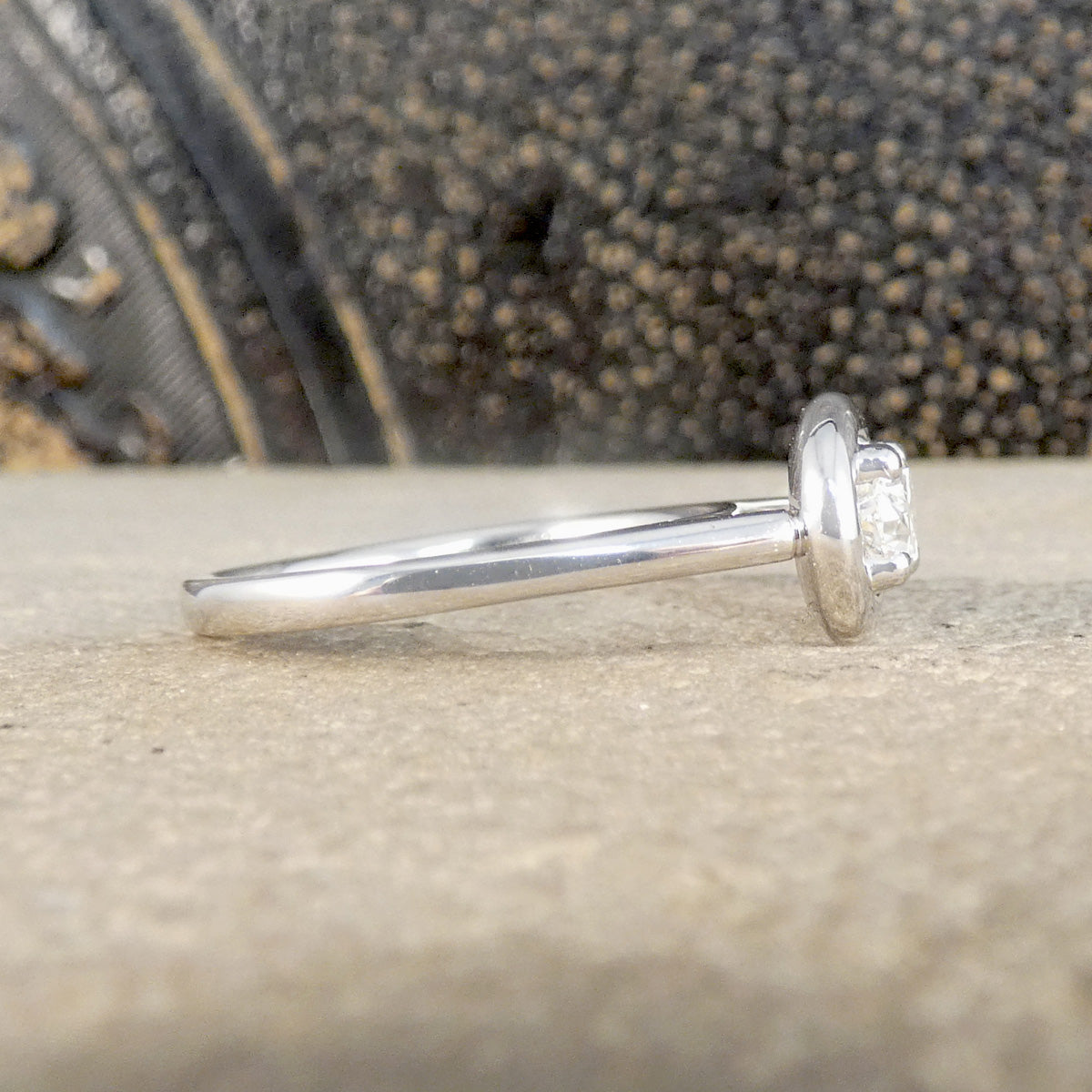 A silver ring with a round bezel setting and clear gemstone, shown in profile on a textured beige stone surface with a dark, blurred background.