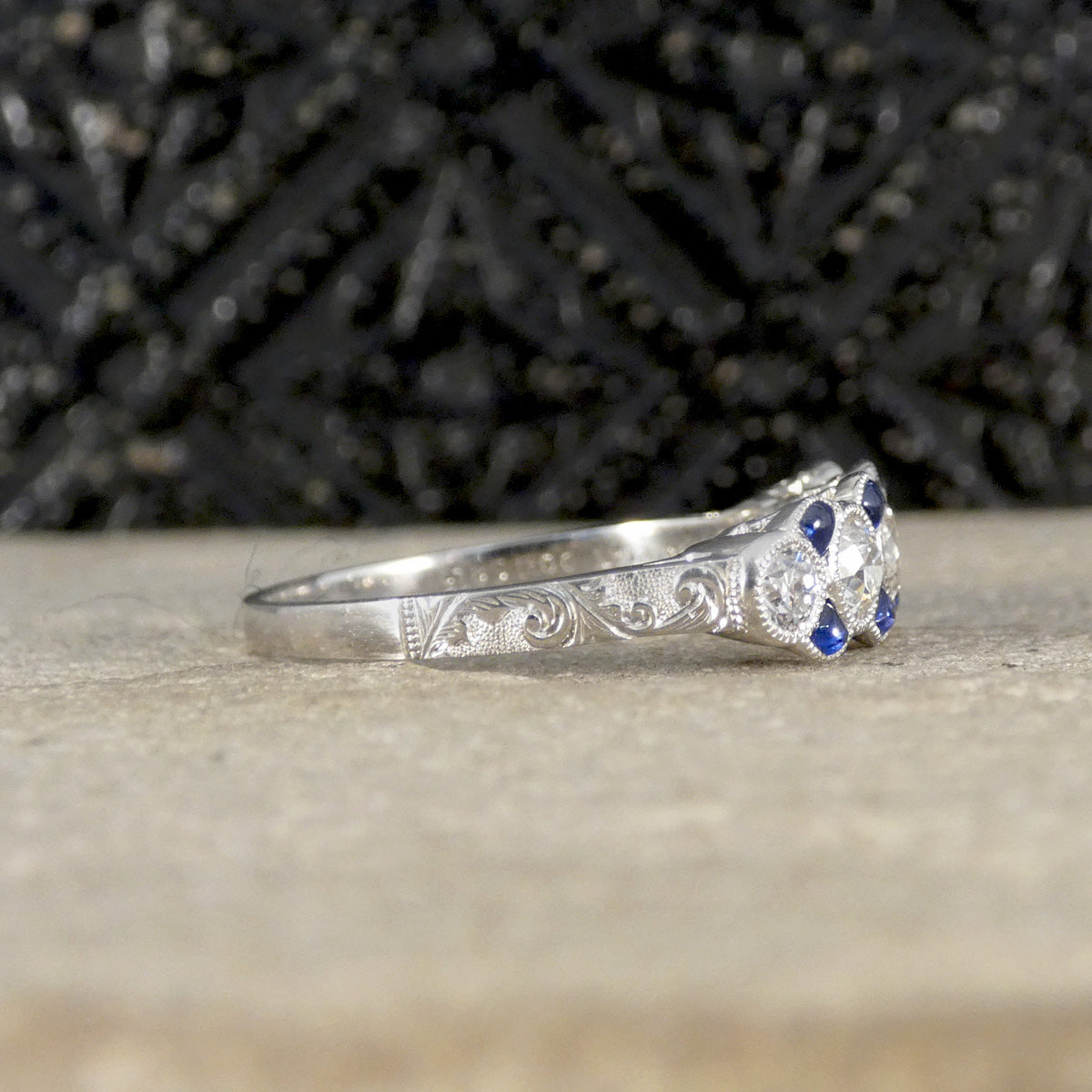 Side view of a vintage-inspired platinum ring featuring intricate hand-engraved scrollwork along the band and a cluster of old-cut diamonds with blue sapphire accents in a milgrain setting.