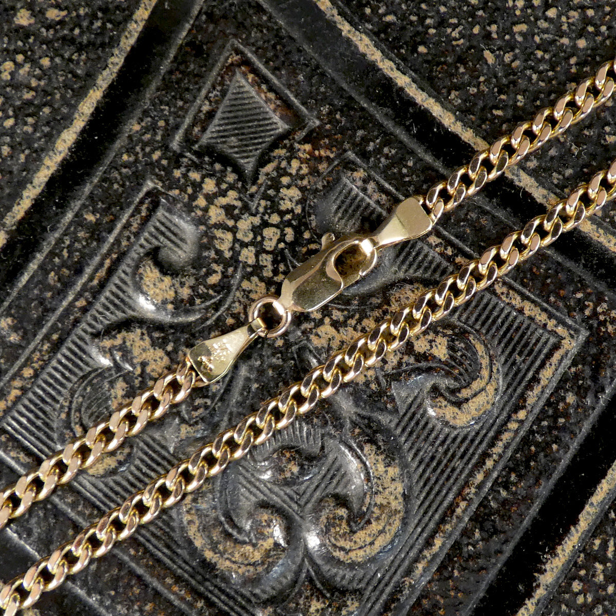 A close-up of a gold-toned metal chain with a lobster clasp resting on a dark, textured surface with embossed decorative patterns.