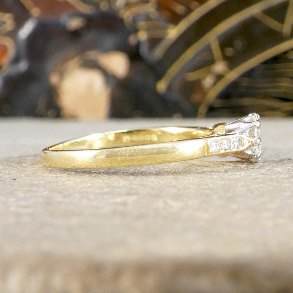 A yellow gold ring with a clear diamond gemstone and small side stones, resting on a textured beige surface with a blurred dark background.