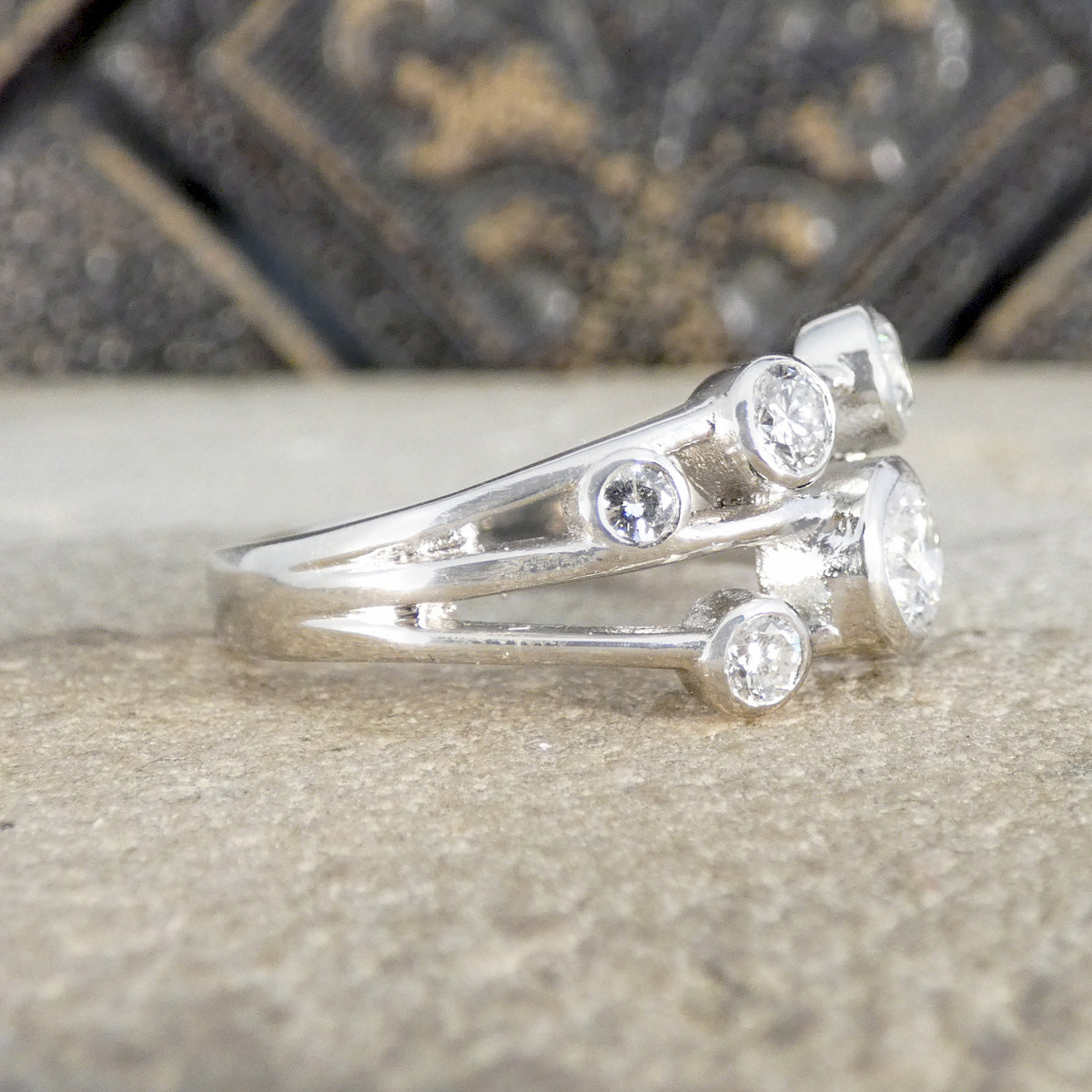A silver-toned ring with three clear round-cut gemstones, photographed close-up on a textured stone surface with a blurred dark decorative background.