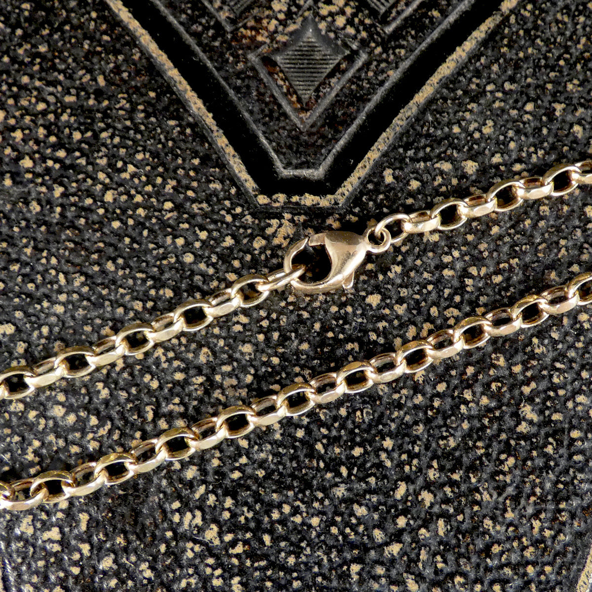 A close-up of a gold chain necklace with a lobster clasp, laid on a textured black and gold patterned surface.