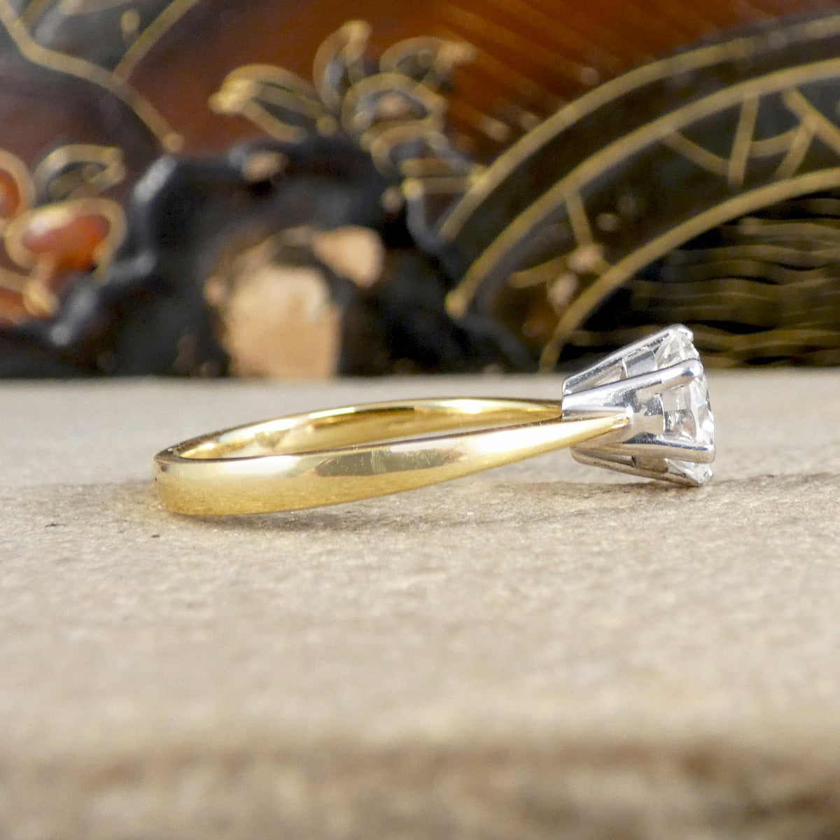 A classic solitaire engagement ring featuring a round brilliant-cut diamond set in a silver-colored stone setting on a yellow gold band, resting on a textured beige surface with a blurred decorative background.