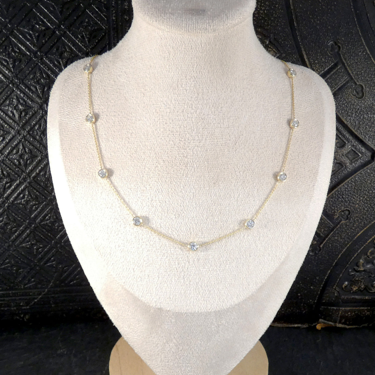 A delicate gold chain necklace with small clear round gemstones spaced along the chain, displayed on a white fabric jewelry bust against a dark textured background.
