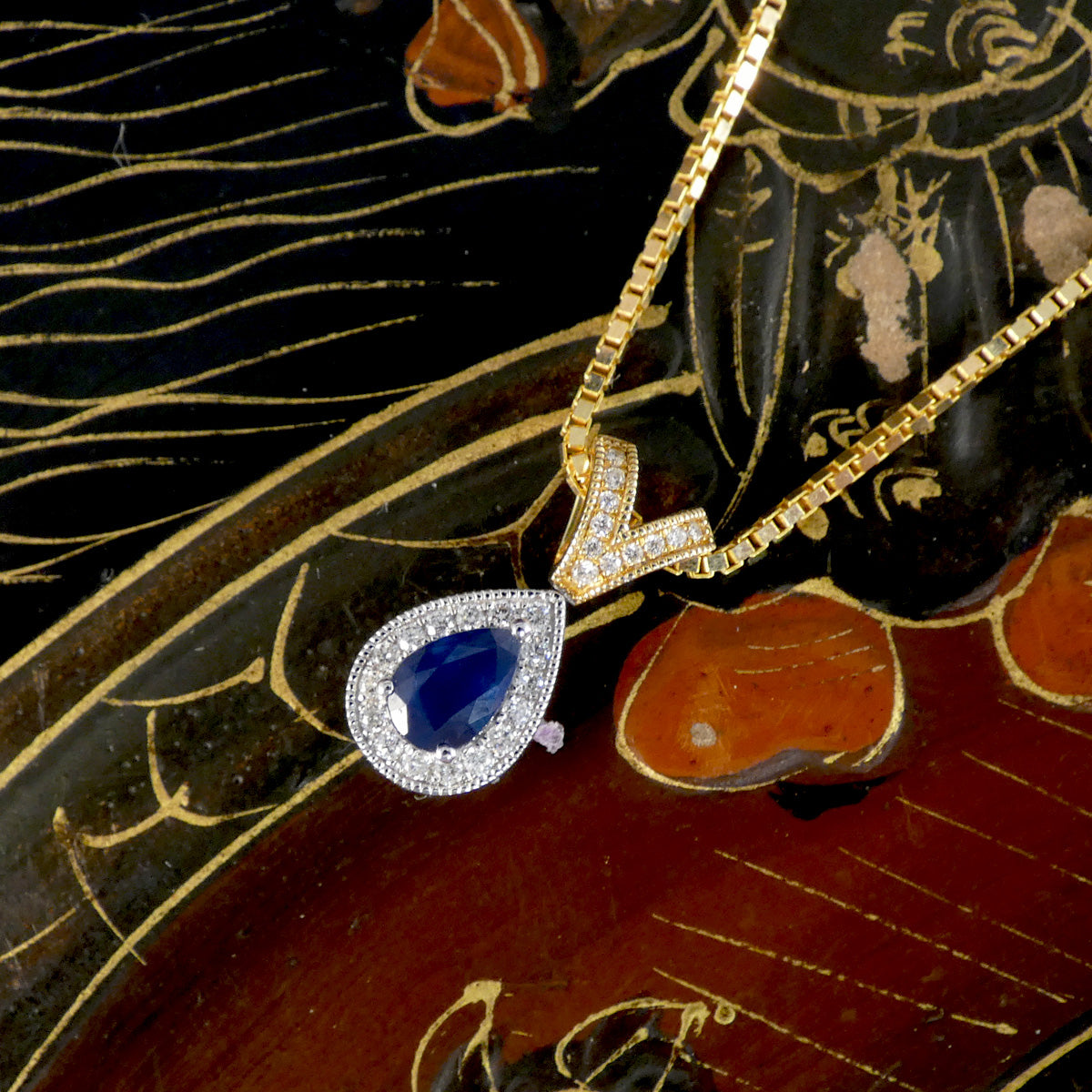 A gold pendant necklace featuring a teardrop-shaped blue sapphire center stone surrounded by a halo of small clear diamonds, displayed on a dark patterned surface.