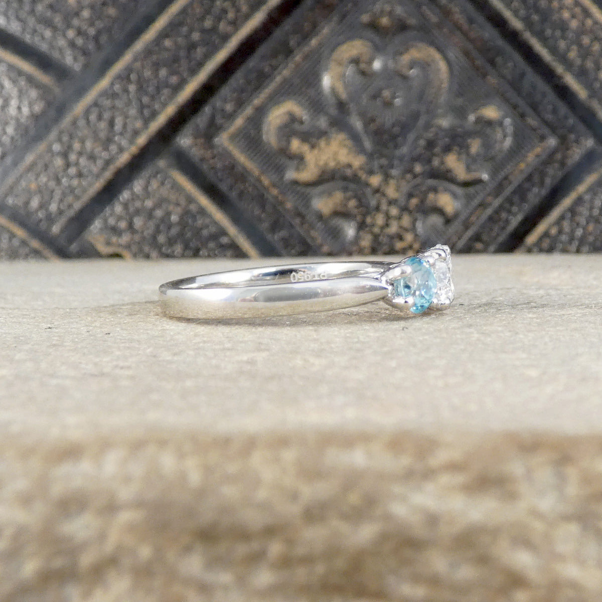A platinum three-stone ring featuring a round brilliant-cut center diamond flanked by two oval light blue zircons, set in a platinum band resting on a textured beige stone surface with a dark patterned tile background.