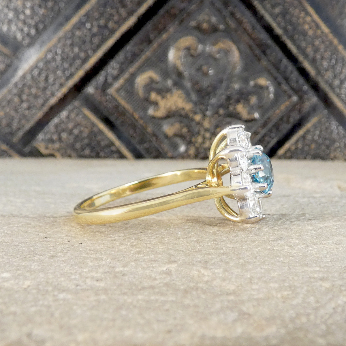 A yellow gold ring with a halo setting featuring a light blue gemstone and small clear accent stones, resting on a textured stone surface with a dark decorative background.