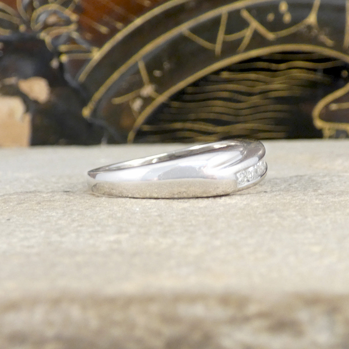 Silver ring with small diamonds on a textured surface with a decorative object in the background