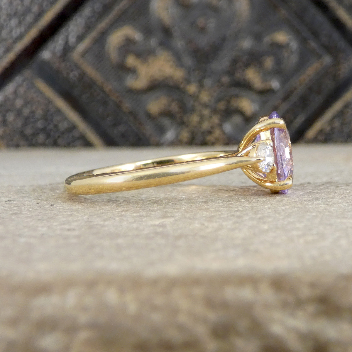 A yellow gold ring with a oval-cut light purple gemstone, resting on a textured stone surface with a dark decorative background.