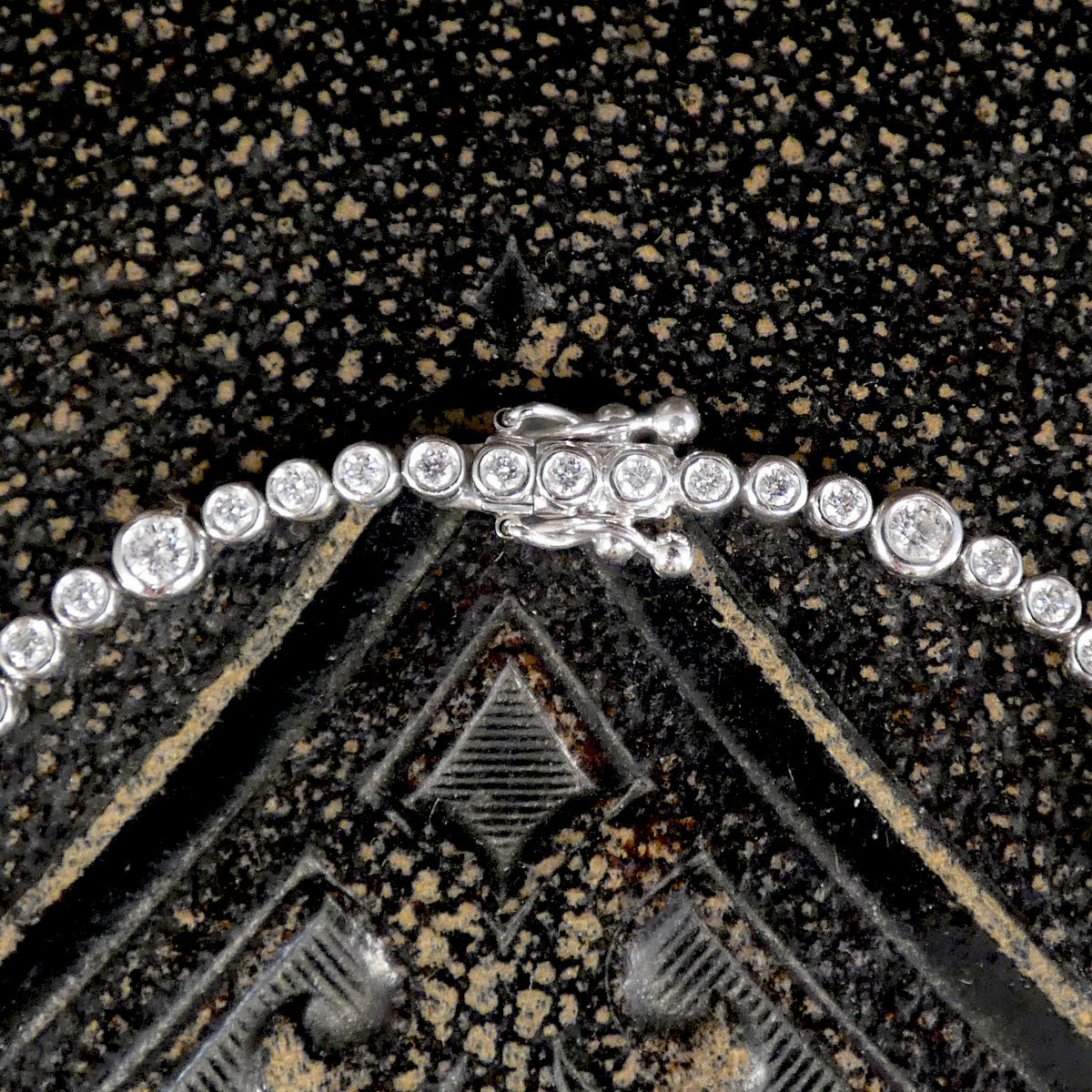 A diamond tennis bracelet with bezel set diamonds of different sizes in white metal on a dark background. Showing snap lock and figure eight double clasp.