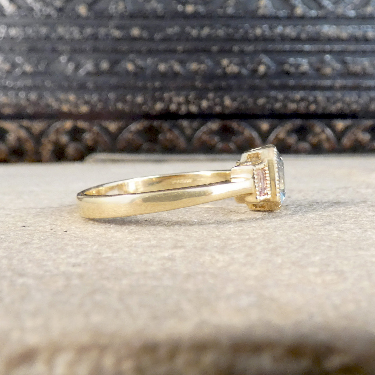 A yellow gold ring with a rectangular gemstone featuring pink and light blue pavé accents, resting on a textured beige stone surface with a dark carved wooden background.