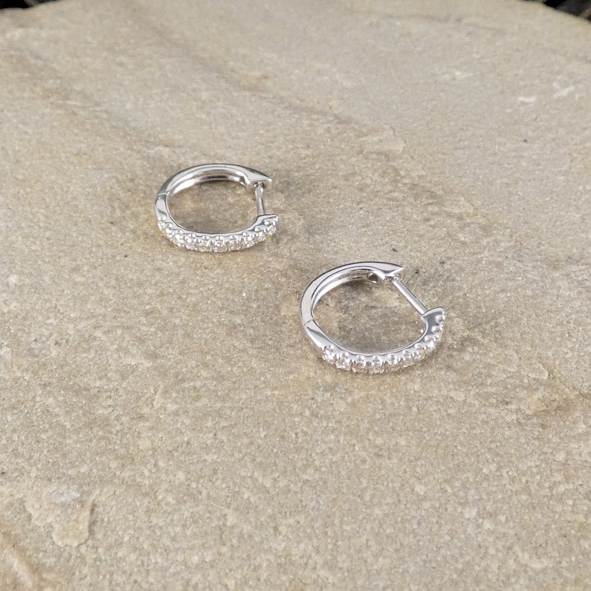 A pair of small white gold huggie hoop earrings with clear pavé stones along the front, displayed on a textured beige stone surface.