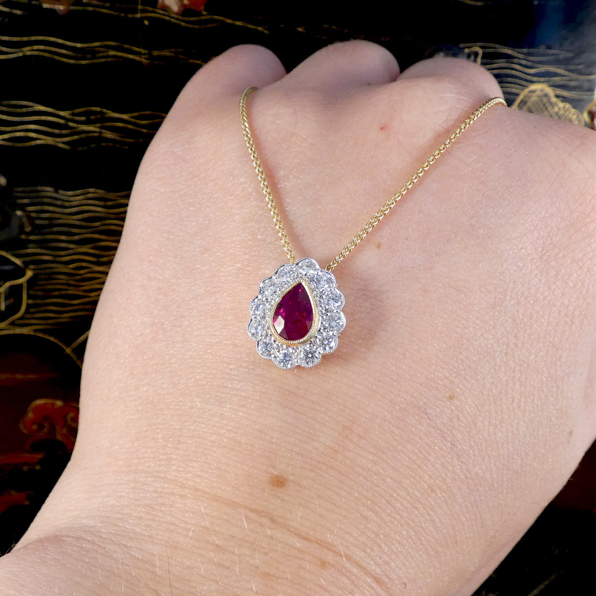Gold necklace with a red ruby gemstone pendant with a diamond surround on a hand against a dark background