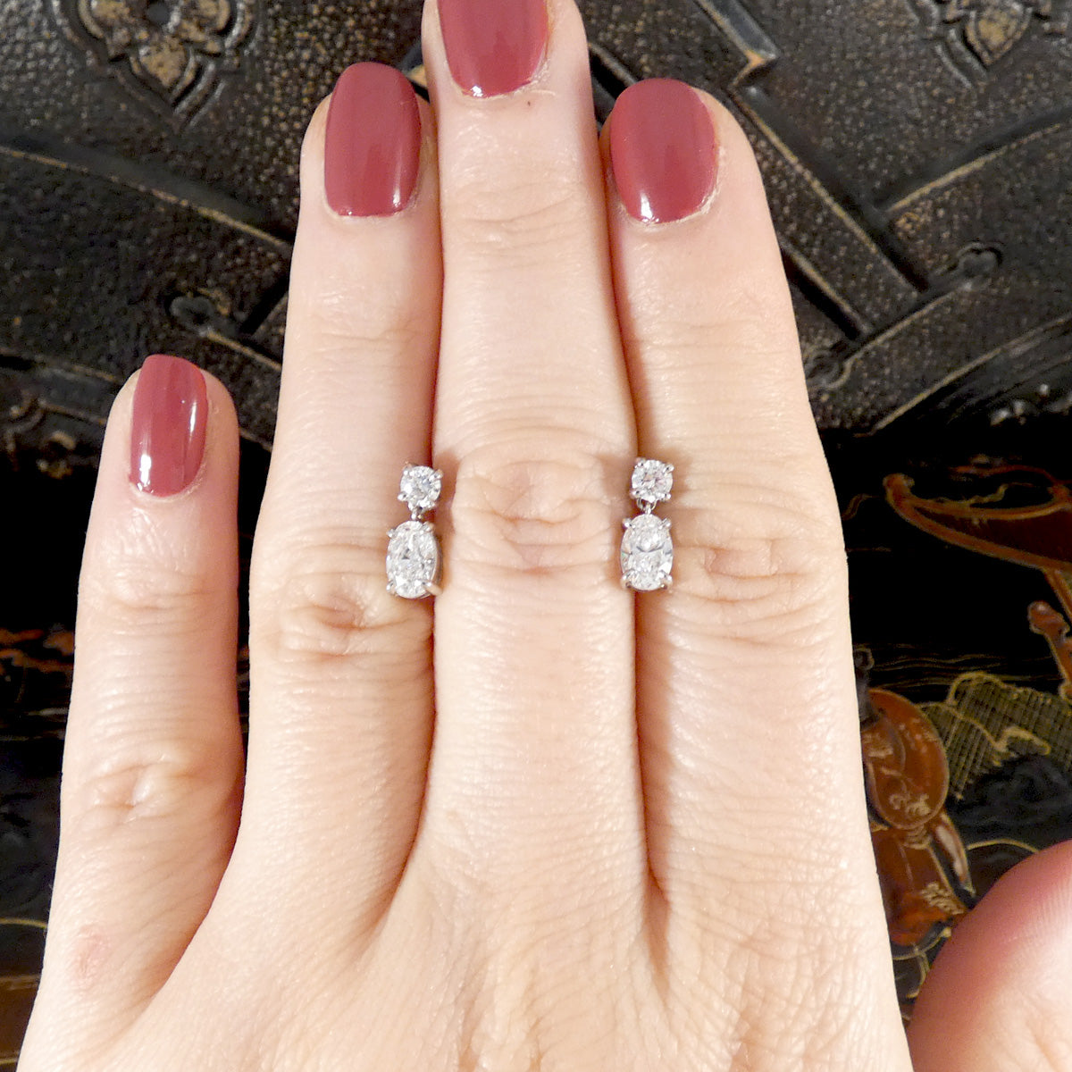 Diamond drop earrings on a hand with pink nail polish against an ornate background