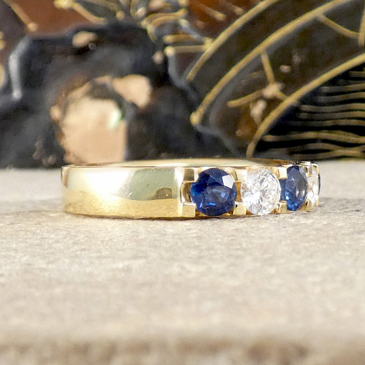 A yellow gold ring featuring three gemstones set in a row: two round-cut deep blue sapphires and one clear round-cut diamond, displayed on a neutral textured surface with a blurred background.