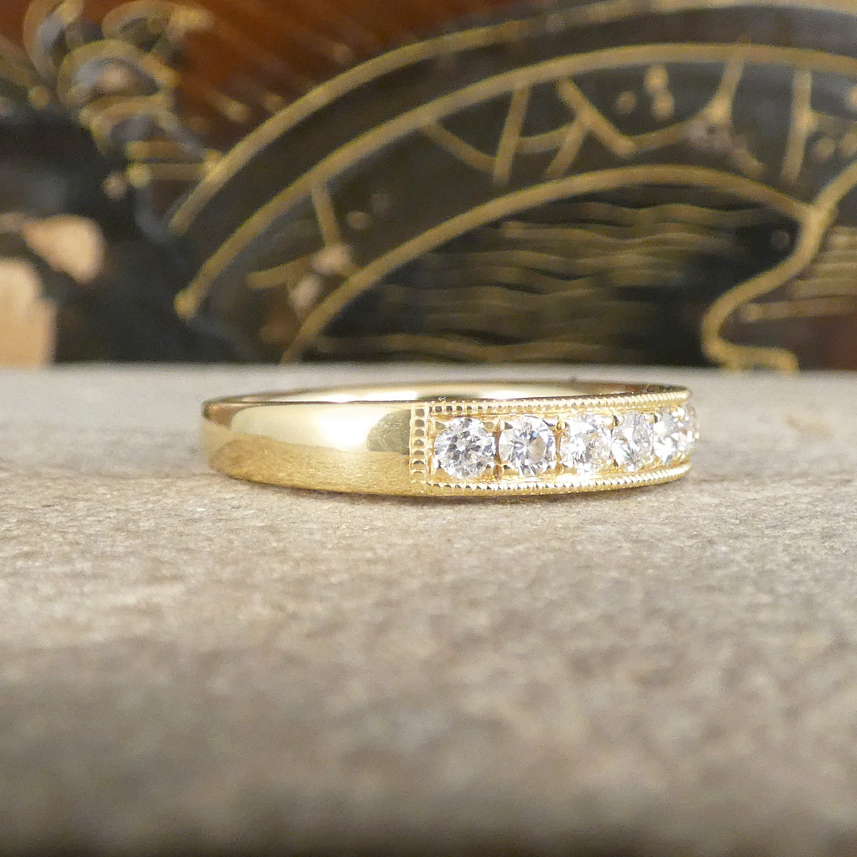 A yellow gold ring with diamond, photographed close-up on a textured stone surface with a blurred decorative background.
