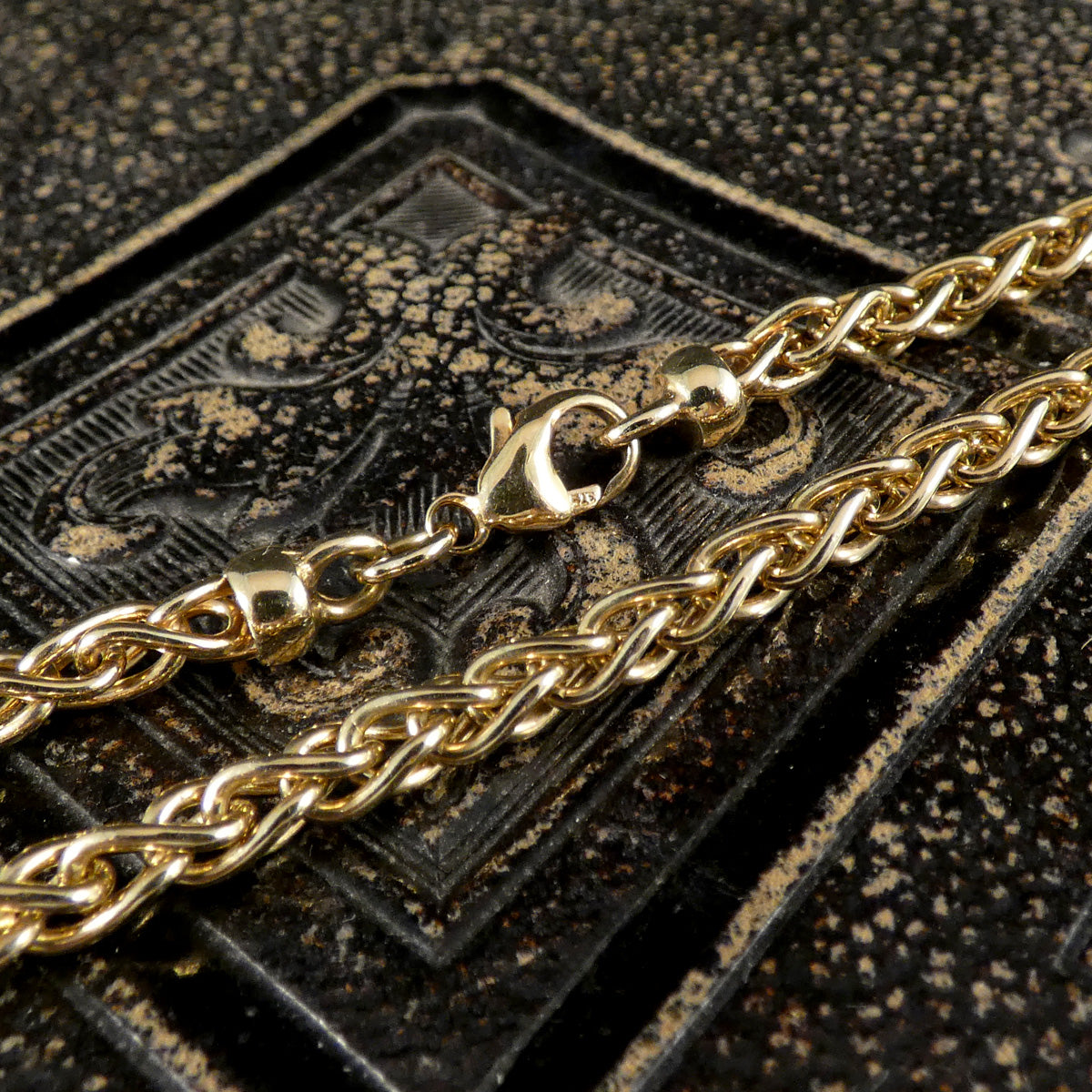 A close-up of a shiny gold chain necklace with a lobster clasp, draped over a dark textured surface.