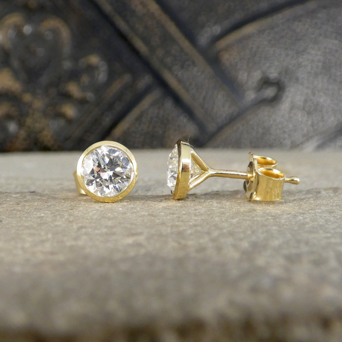 A pair of yellow gold stud earrings with round bezel-set clear gemstones, shown on a textured stone surface with a dark, out-of-focus background.