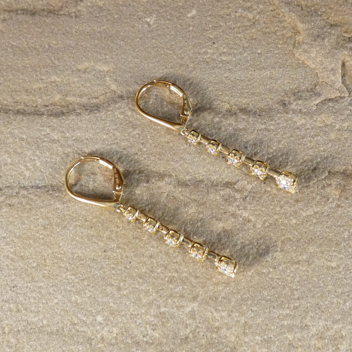 A pair of gold leverback drop earrings with small clear stones, displayed on a textured beige stone surface.