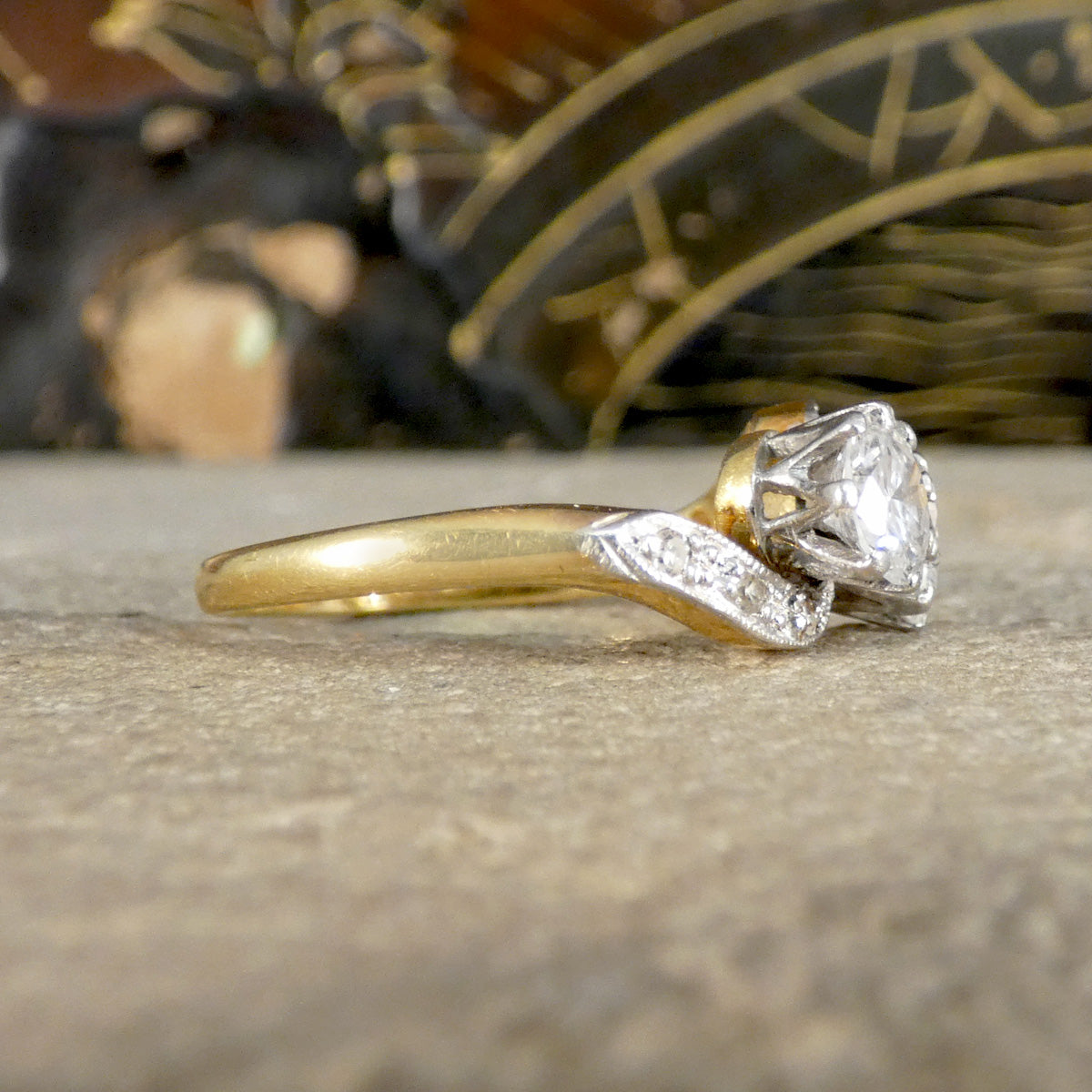 A yellow gold ring with a diamond solitaire and a diamond-accented split shank, resting on a textured stone surface with a blurred decorative background.