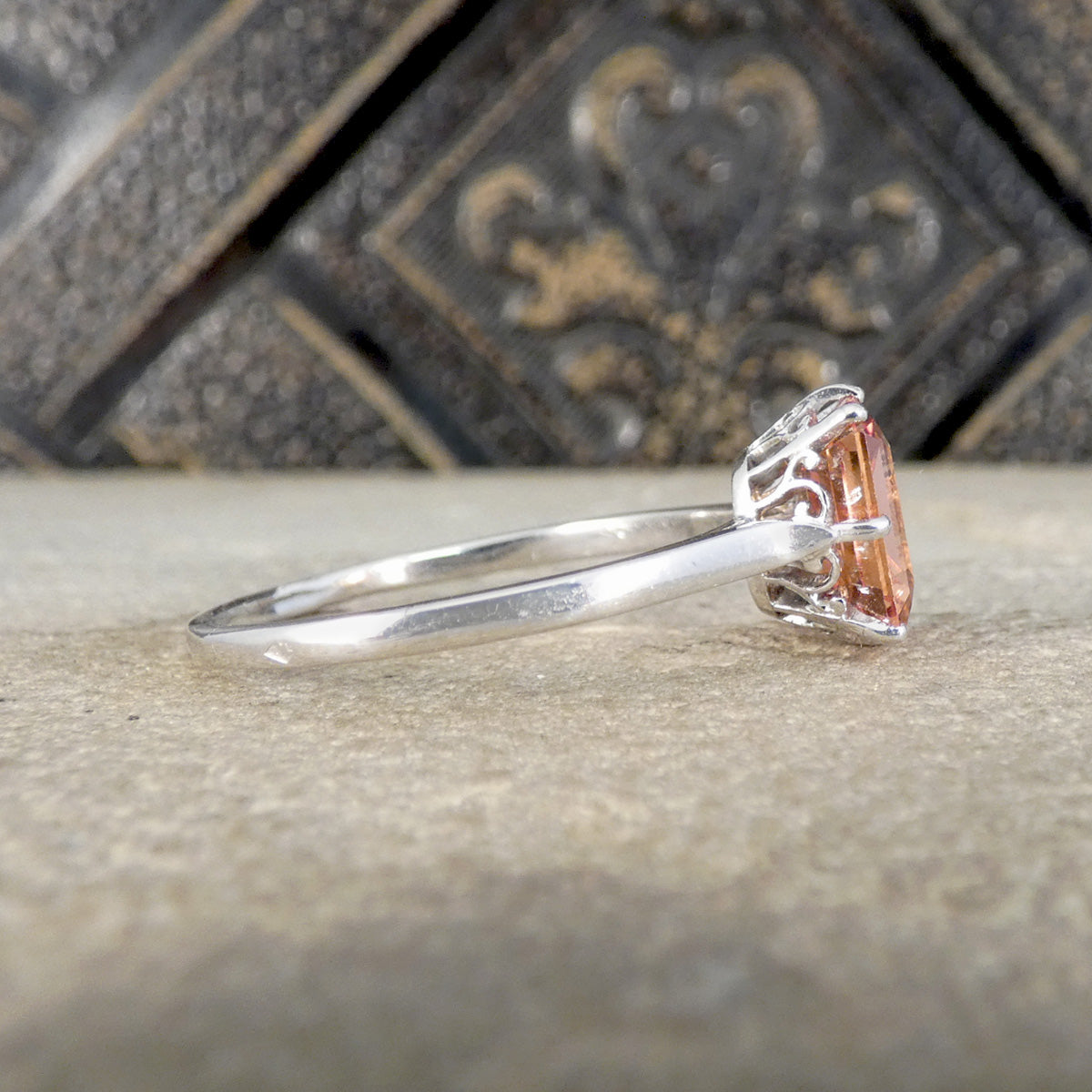 A silver ring featuring a emerald-cut orange sapphire gemstone in a decorative filigree setting, resting on a textured stone surface with a dark, ornate background.