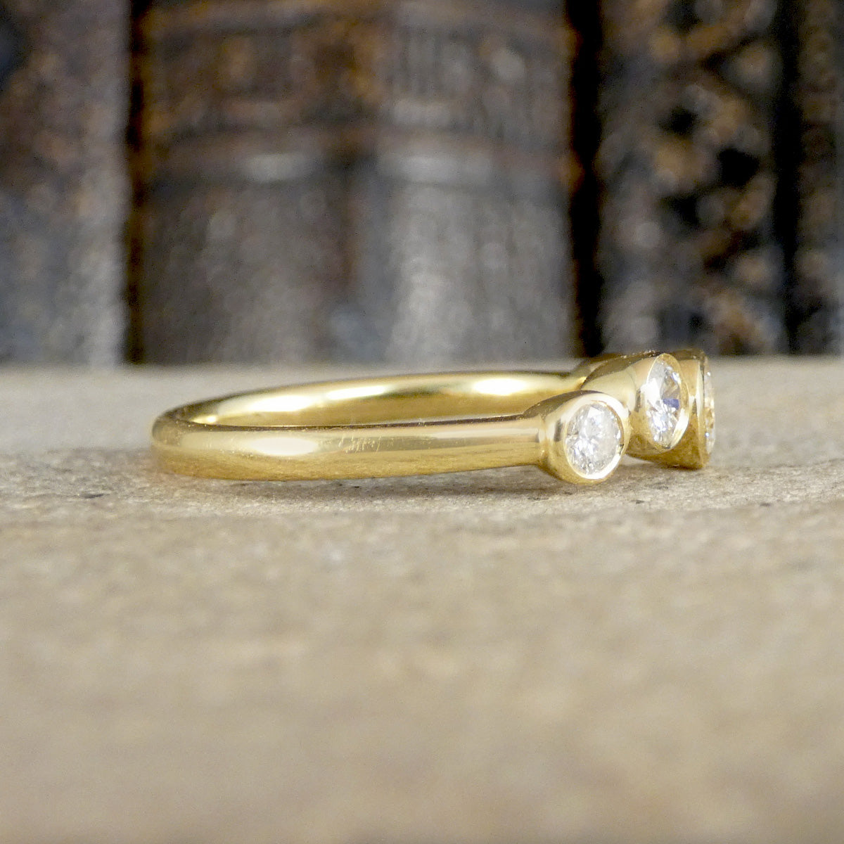 A yellow gold ring with two small clear gemstones, resting on a textured beige stone surface with a blurred dark background.