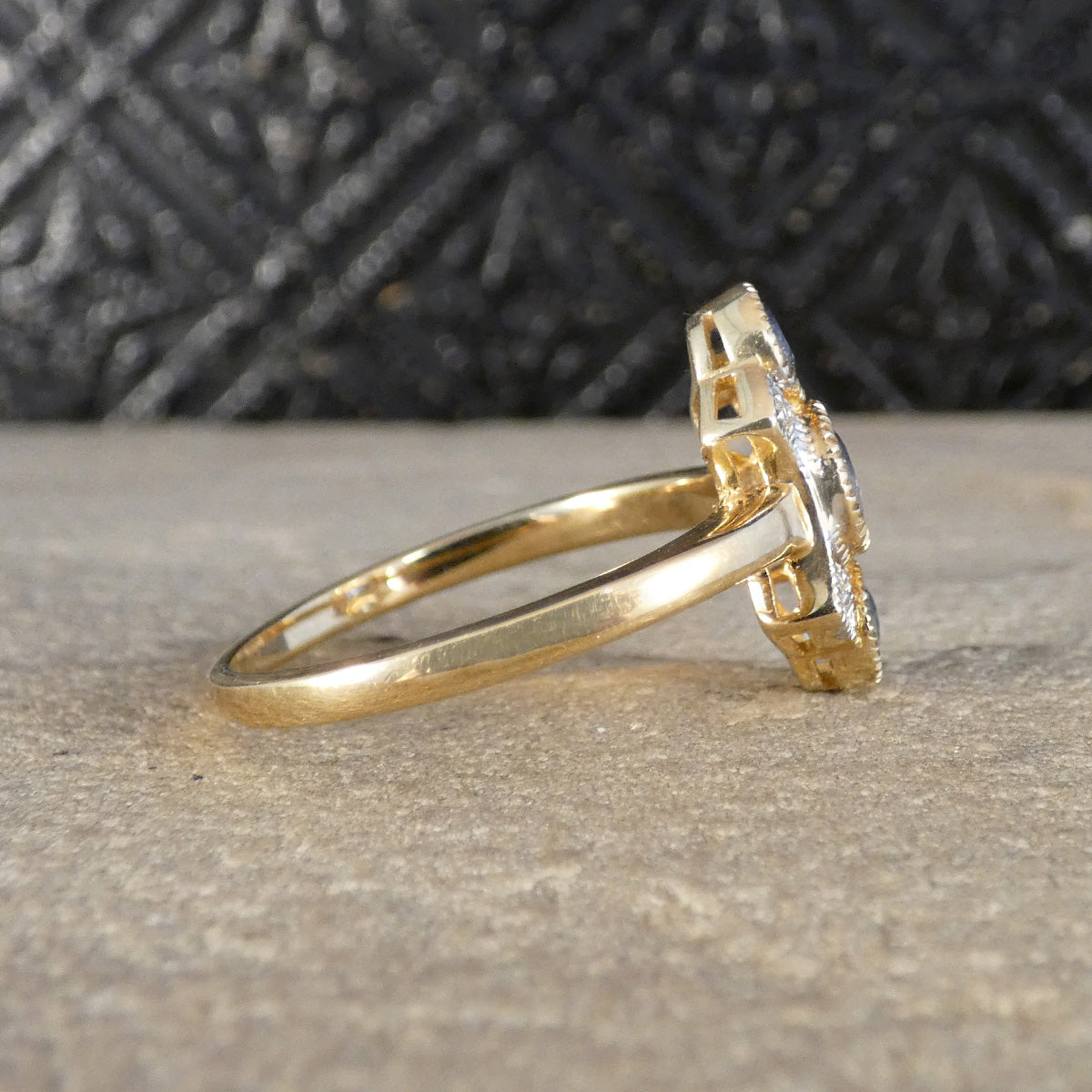 A yellow gold ring with a floral cluster setting, photographed on a textured gray stone surface with a dark, patterned background.