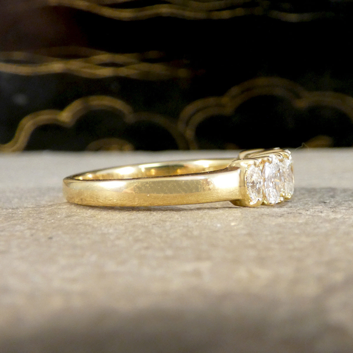 A yellow gold ring with a three-stone setting featuring clear round-cut gemstones, resting on a textured light gray surface with a dark, patterned background.