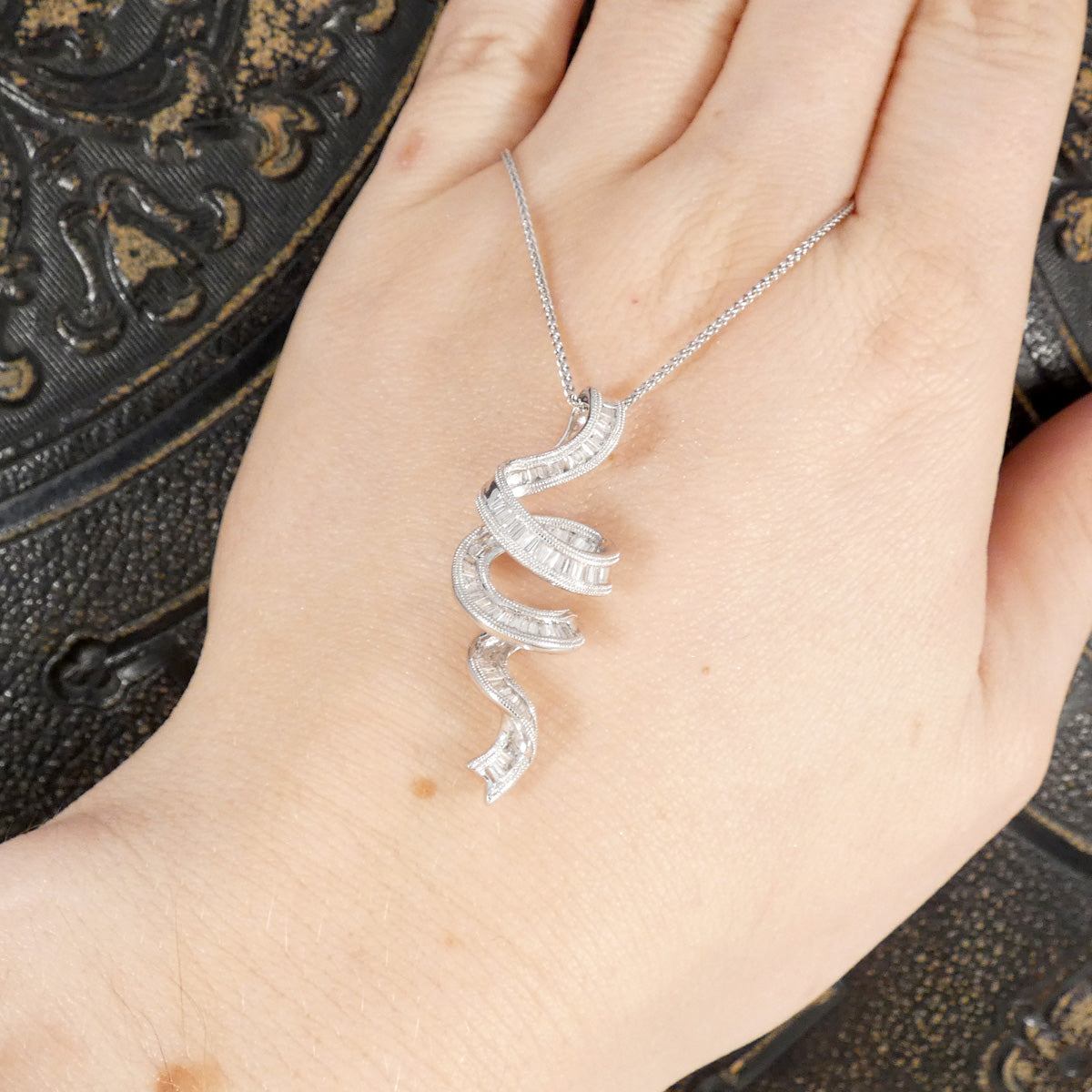Silver ribbon shaped diamond set pendant on a chain held by a hand against a dark background
