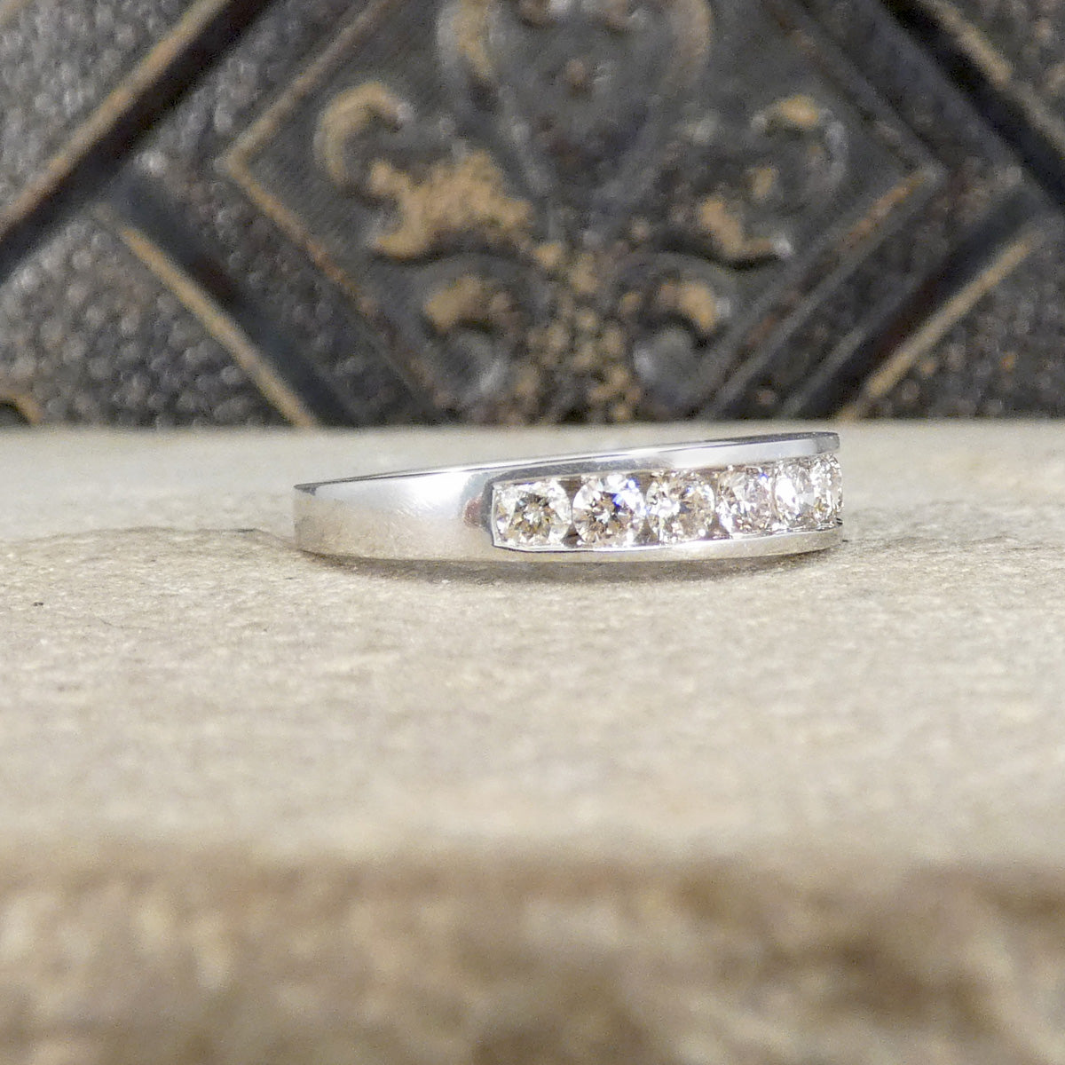 A 9ct white gold band ring with five clear round-cut gemstones set in a row, resting on a textured beige stone surface with a dark, ornate background.