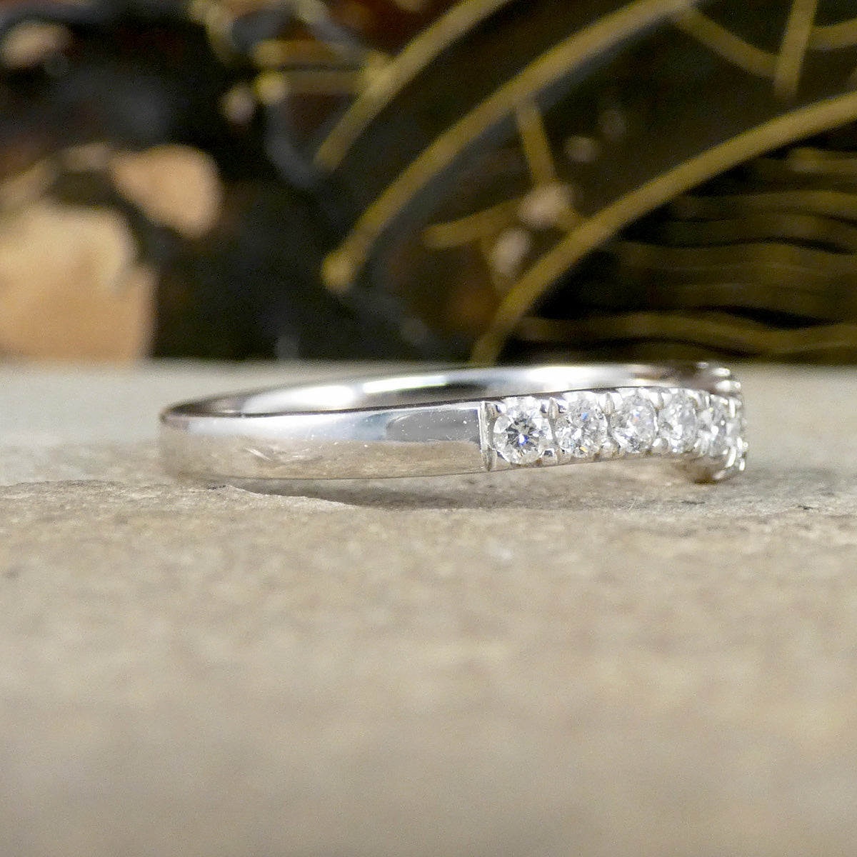 A silver band ring featuring a row of clear round-cut gemstones on the front, photographed close-up on a textured beige surface with a blurred dark background.