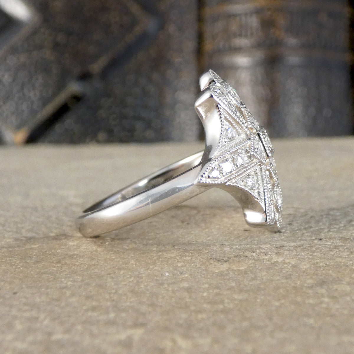 A silver Art Deco style ring with a geometric, fan-like top set with small clear stones, resting on a textured stone surface with a blurred dark background.
