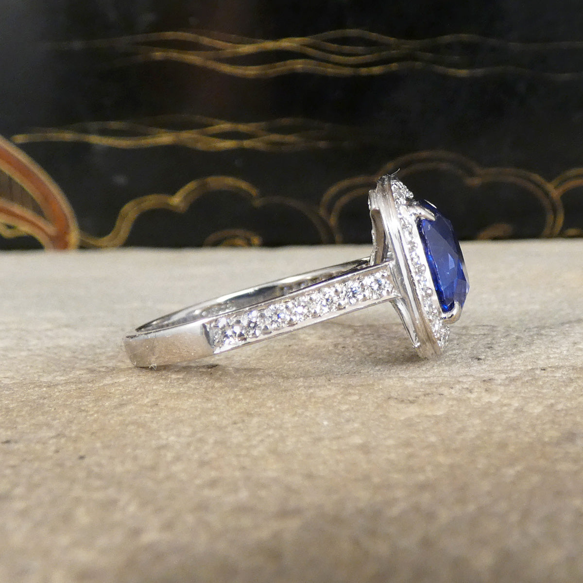 A silver ring featuring a large oval blue sapphire gemstone in a halo setting, with a pavé band, resting on a textured beige stone surface.