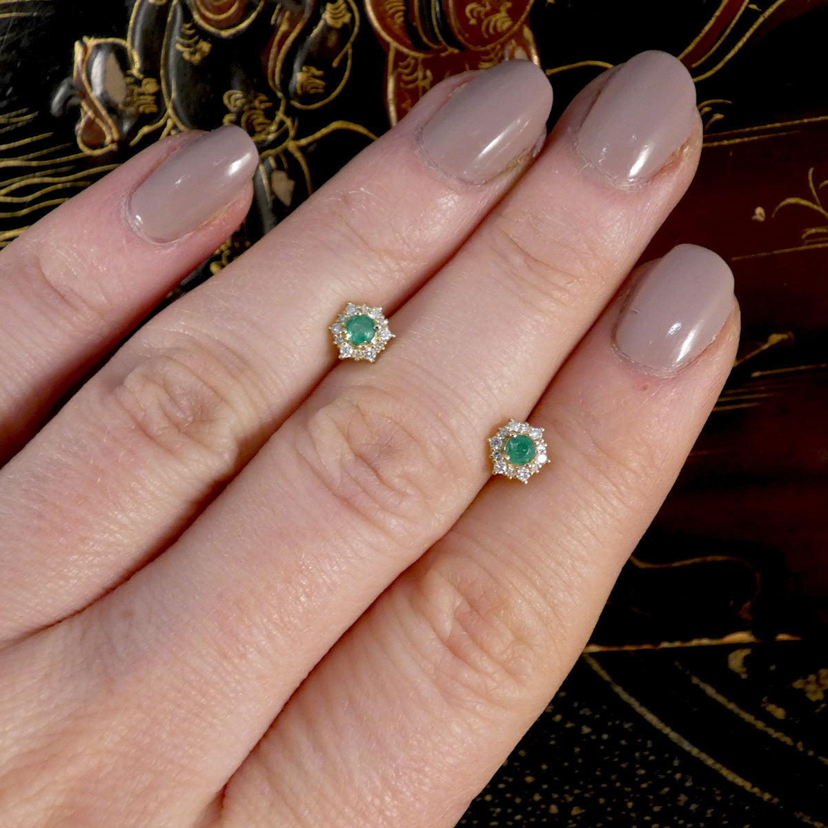 Hand showing a pair of gold earrings with a green gemstone against a decorative background