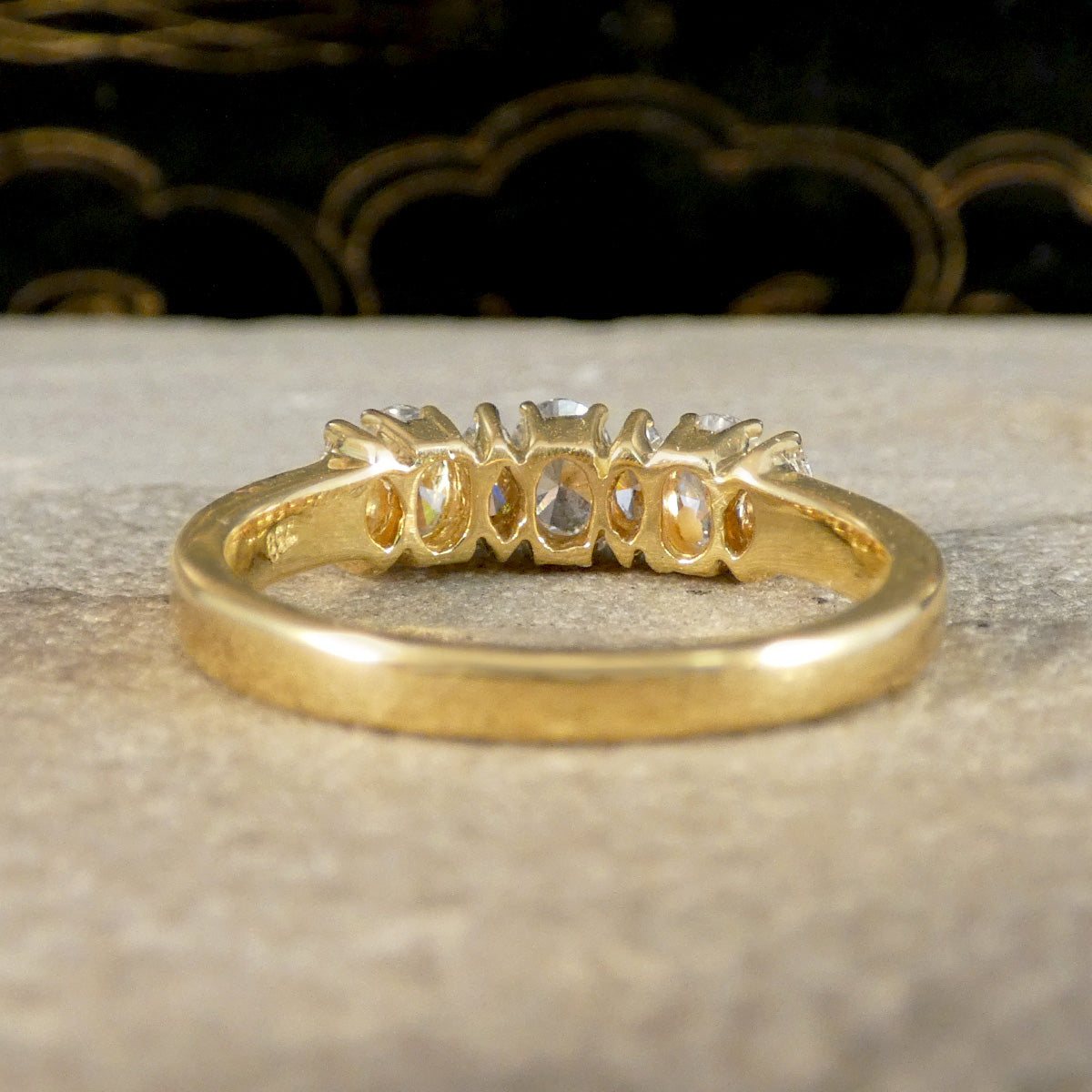 A yellow gold ring with a row of small clear gemstones, photographed close-up on a textured light surface with a dark decorative background.