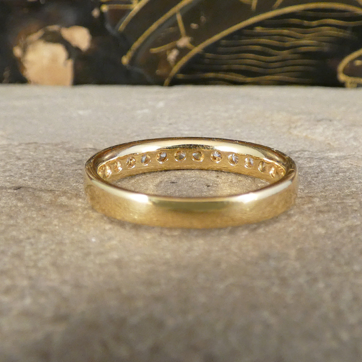 A yellow gold eternity ring with a row of small clear stones set along the band, resting on a textured gray stone surface.