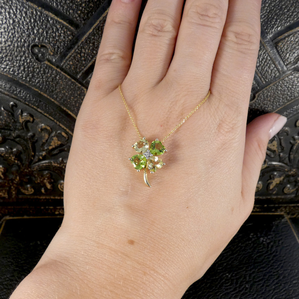 A gold necklace with a four-leaf clover pendant featuring green gemstones and a clear center stone, shown worn on a person's hand against a dark textured background.