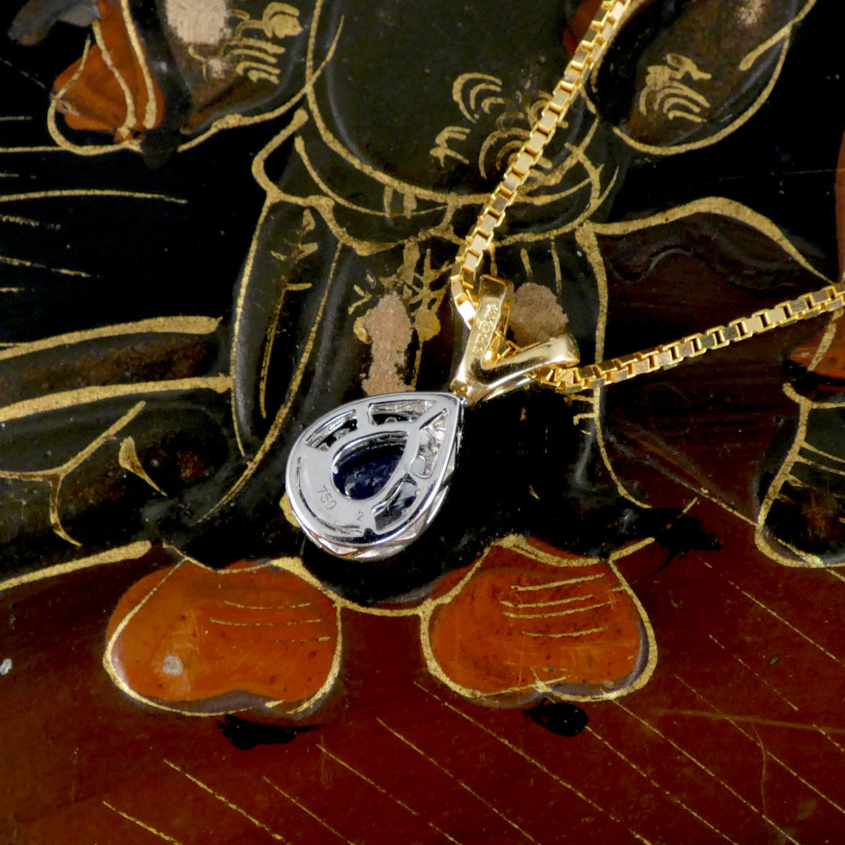 A close-up of a gold chain necklace with a silver-toned pendant featuring a dark blue inlay, resting on a black and gold decorative wooden surface.