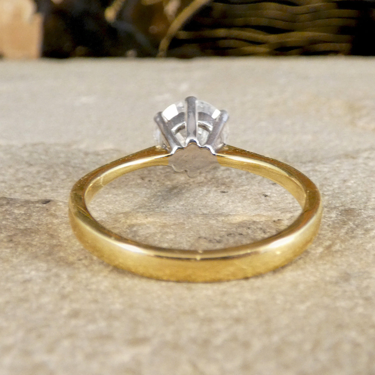 A yellow gold ring band with a silver-toned six-prong setting holding a clear gemstone, resting on a textured beige stone surface.