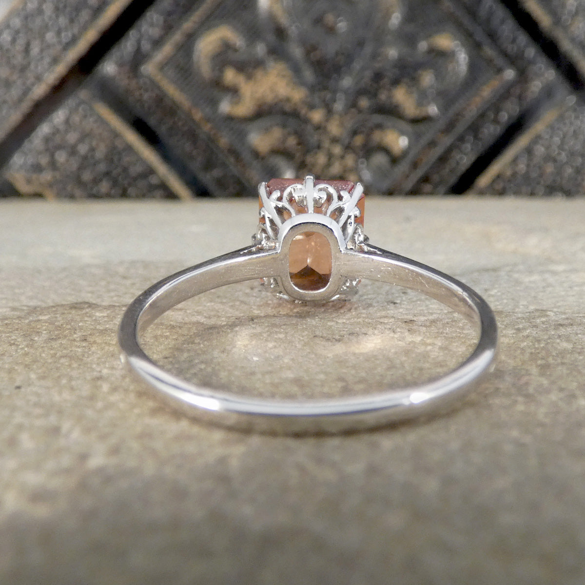 A silver ring with an oval peach-colored gemstone and a decorative crown-like top, resting on a textured stone surface with a dark ornate background.