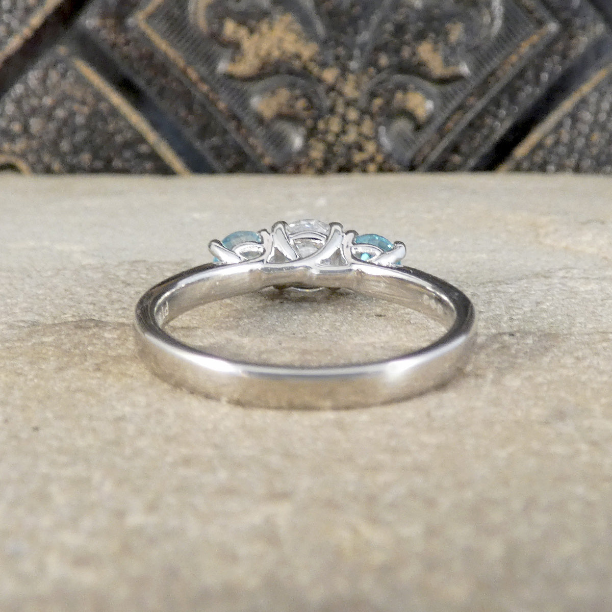 A silver ring with a three-stone setting featuring a central clear round diamond and two small light blue gemstones on either side, resting on a textured beige stone surface.