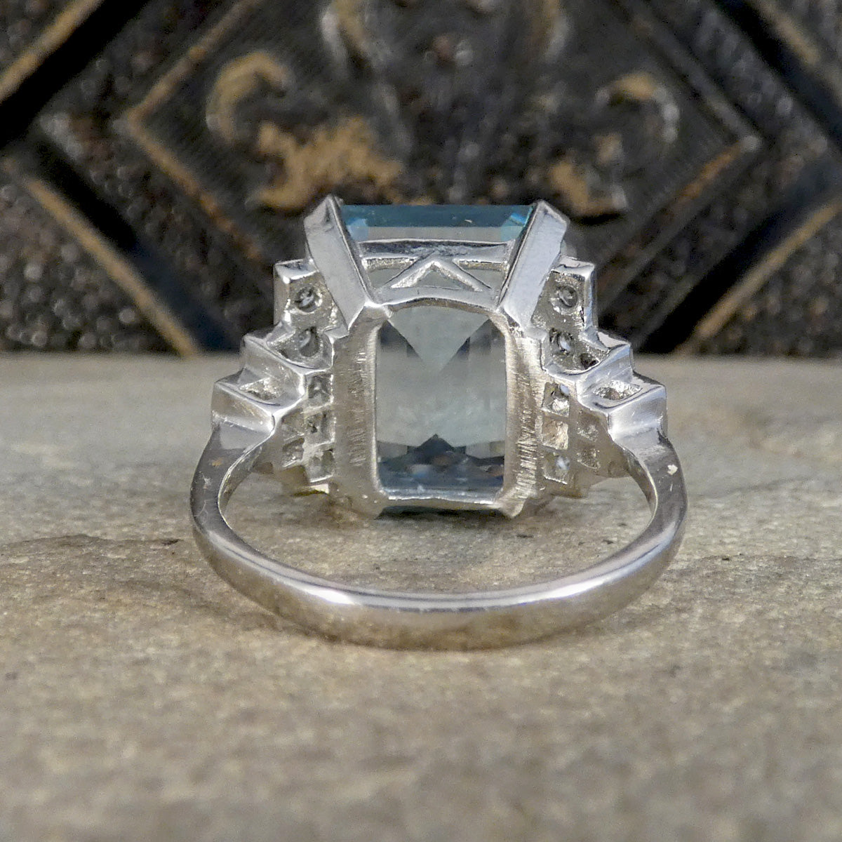 A platinum ring featuring a rectangular light blue gemstone in a geometric setting, flanked by smaller clear stones and decorative silver elements, resting on a textured stone surface.