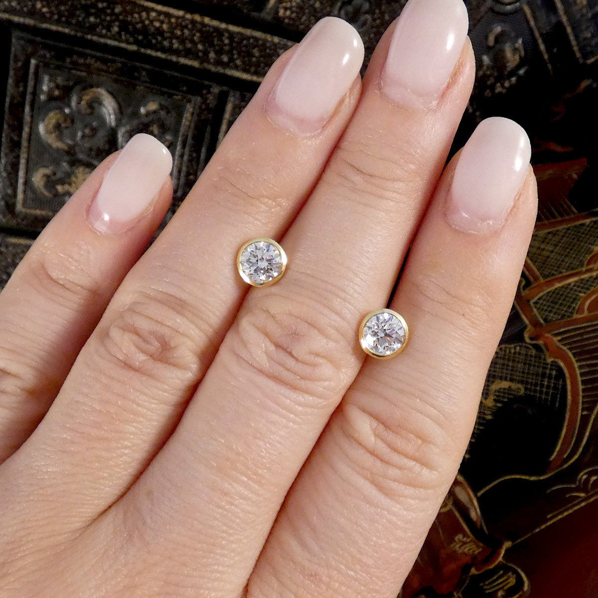 A close-up of a hand with pale pink manicured nails wearing two round stud earrings with gold settings and clear gemstones, against a dark ornate background.