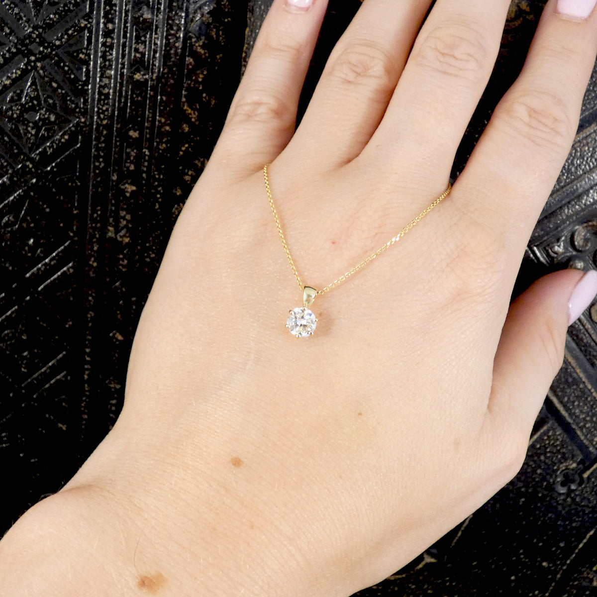 A gold necklace with a single round clear diamond pendant, shown worn on a person's hand against a dark textured background.