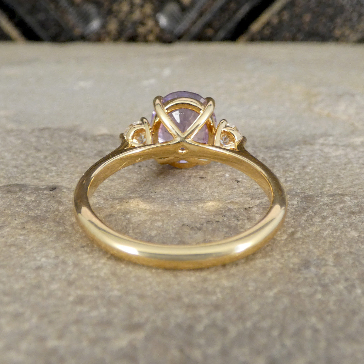 A gold ring with a round light purple gemstone, resting on a textured gray stone surface.
