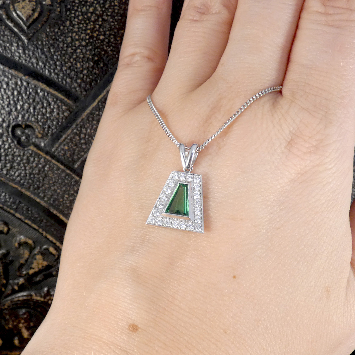 A silver-toned geometric pendant necklace with a green triangular center stone and a halo of small clear stones, shown worn on a person's hand.